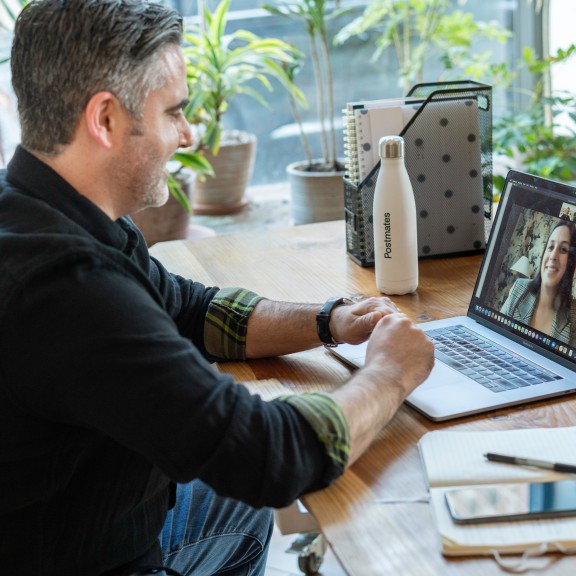 Man is setting at a table with his laptop open. On the screen is a woman. They seem to be in a meeting because he has a notebook, pen, water bottle, files, and his phone on the tabletop around him.