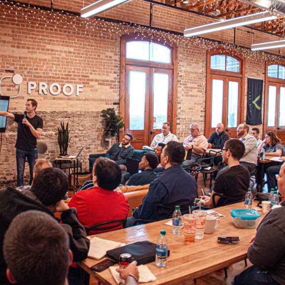 Man in black t-shirt giving a presentation to a group of staff in an office. He holds a microphone and points at a screen..