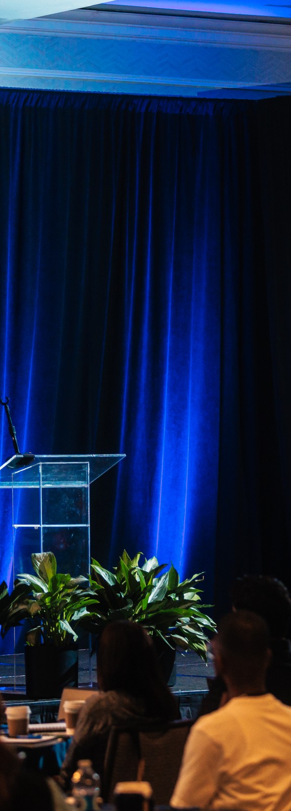 Female on stage speaking to a large crowd with a PowerPoint slide with graphics showing