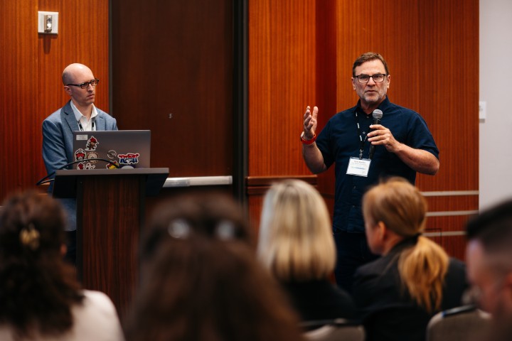 Two presenters with audience, one at podium, other talking on microphone.