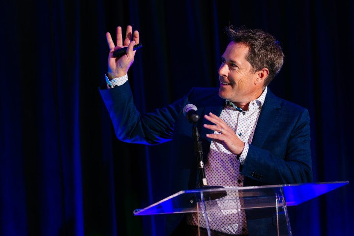 Man on stage with hand up and microphone/podium