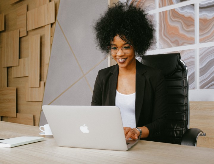 A woman is sitting at her desk with an apple laptop in front of her and wooden abstract walls behind her. She wears a blazer and smiles.