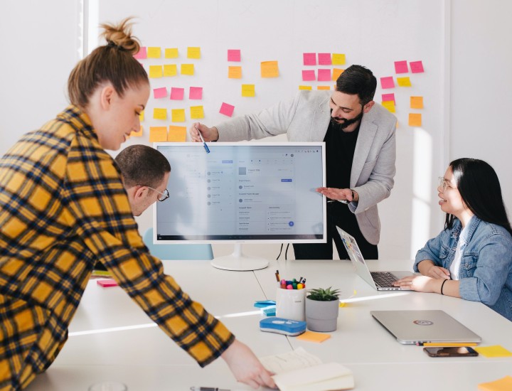 A man is standing beside a PC screen pointing to it as he is presenting. There are two people sitting at the meeting table looking at him and his presentation. There is another person in a flannel grabbing files from the table.