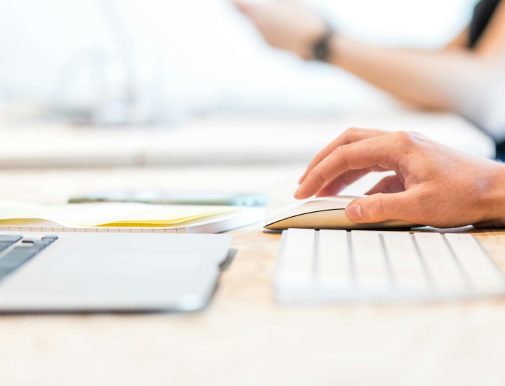 Hand on mouse with keyboard and laptop in front. Blurred arms holding something in the background.
