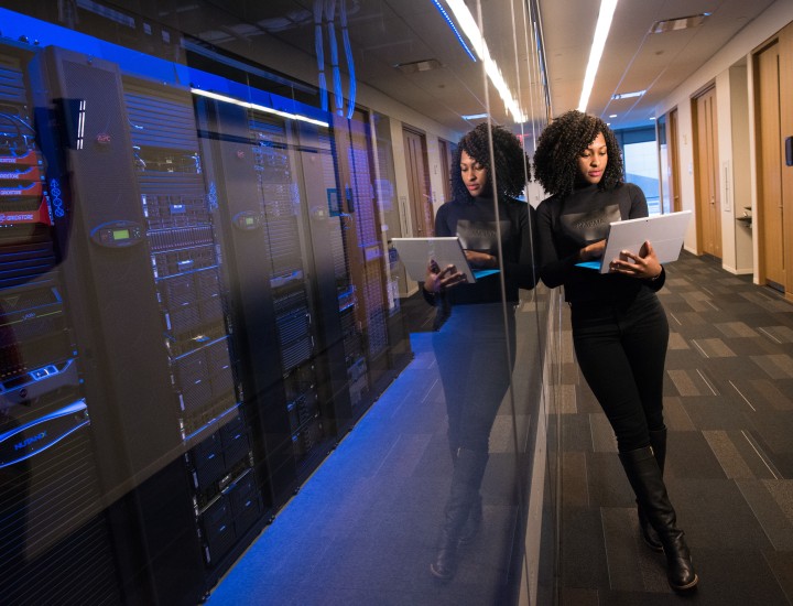 Woman leaning against windows of server room while she is on laptop.