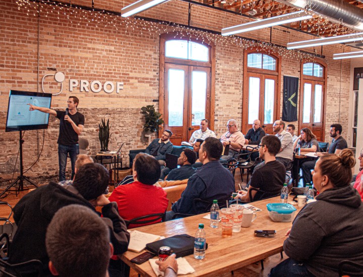 Man in black t-shirt giving a presentation to a group of staff in an office. He holds a microphone and points at a screen.. 